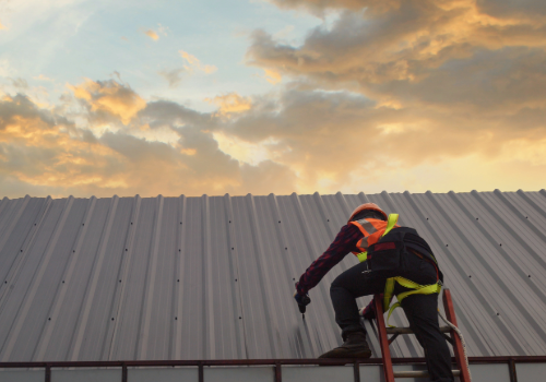 Roofer in high-visibility gear using a tool to repair a metal roof on a commercial building during a sunset.