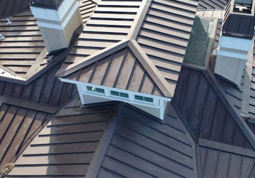 Aerial view of a complex roof with standing seam steel panels, featuring multiple chimneys and geometric shapes.